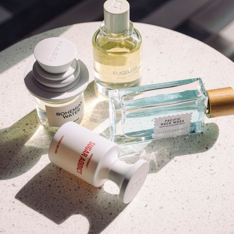 Cuatro frascos de perfume diferentes sobre una superficie blanca moteada, iluminados por la luz natural del sol, cada uno con formas y etiquetas únicas.