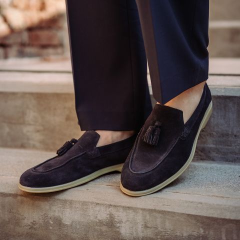 Persona con mocasines de borlas de ante azul marino oscuro con suelas color crema, combinados con pantalones azul marino, de pie sobre escalones de piedra.