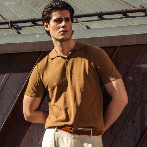 Un hombre con un polo marrón y pantalones beige está de pie al aire libre a la luz del sol, con una mano en el bolsillo, frente a una estructura metálica.