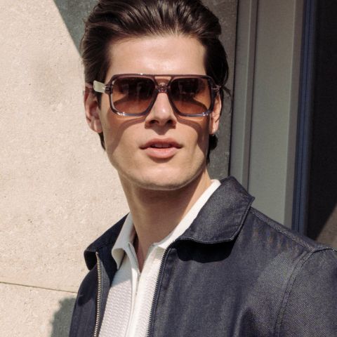 Un hombre con unas grandes gafas de sol marrones y una chaqueta oscura está de pie al aire libre a la luz del sol y mira ligeramente hacia un lado.