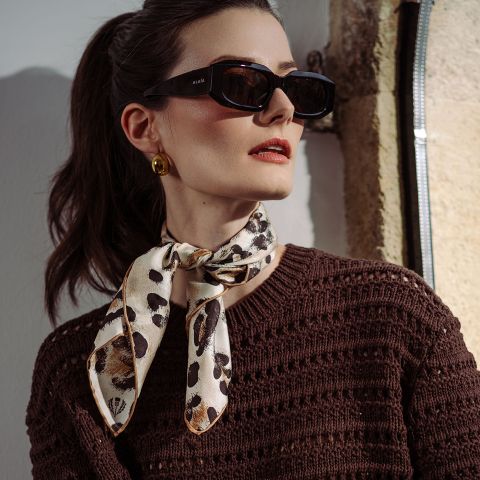 Mujer con gafas de sol negras, pendientes dorados, un top de punto marrón y una bufanda estampada atada al cuello, de pie a la luz del sol.