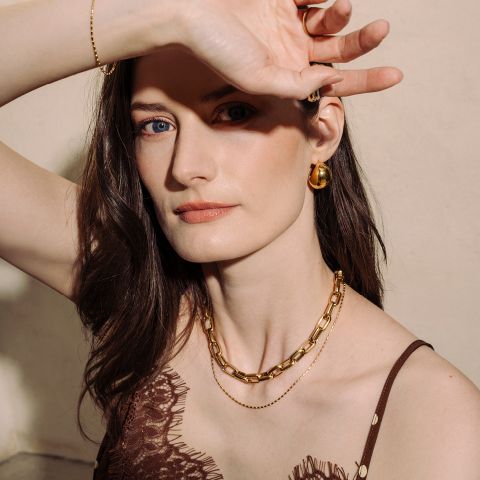 Una mujer de piel clara y cabello castaño, que lleva joyas de oro, posa con un brazo levantado y mira a la cámara con una suave luz natural.