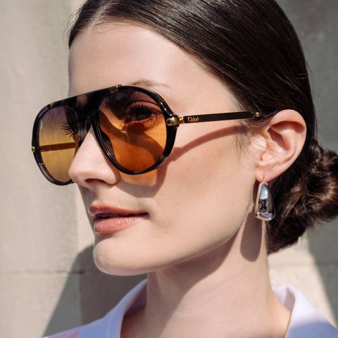 Mujer de cabello oscuro recogido en un moño bajo, con gafas de sol marrones de gran tamaño y pendientes plateados, de pie frente a una pared clara.