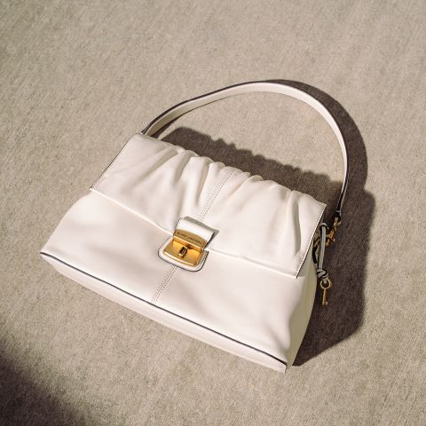 Bolso color crema con hebilla dorada y asa sencilla, apoyado sobre una superficie de hormigón claro a la luz natural del sol.