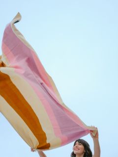 Una mujer sonríe y sostiene una manta grande de rayas de colores frente a un cielo azul claro.