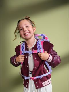 Una joven sonriente con una chaqueta marrón y un vestido blanco, que lleva una mochila colorida, está de pie frente a un fondo verde.