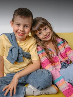 Un niño sonriente y una niña sonriente se sientan juntos en un sofá amarillo. El niño lleva una camisa amarilla, la niña un jersey de rayas de colores.