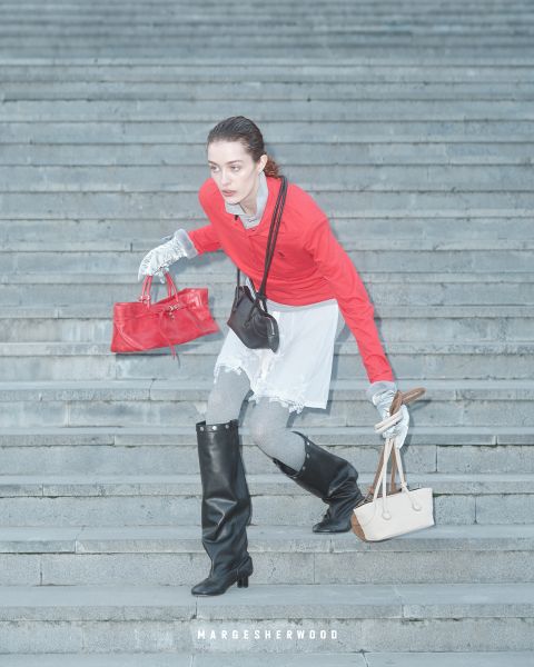 Una mujer con una blusa roja, guantes grises y botas negras baja una escalera de piedra, con un bolso rojo y uno beige en la mano, debajo el texto "Margesherwood".
