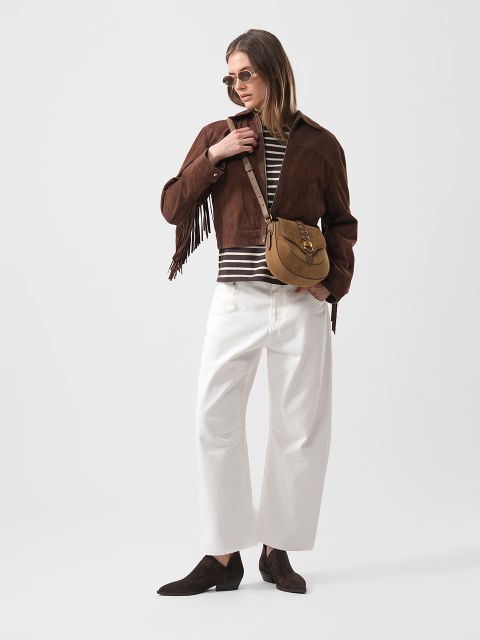 Mujer con chaqueta de ante marrón con flecos, camisa a rayas, pantalón blanco de pierna ancha, zapatos marrones, gafas de sol y bolso bandolera marrón claro, de pie con confianza.