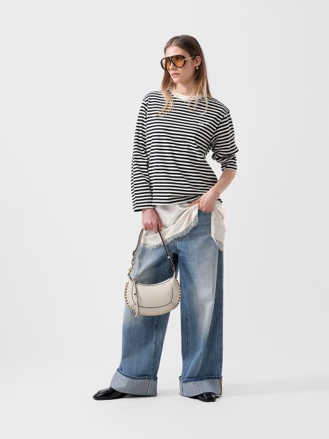 Mujer con un jersey de rayas blancas y negras, vaqueros de pierna ancha, gafas de sol y bolso blanco delante de un sencillo fondo blanco.