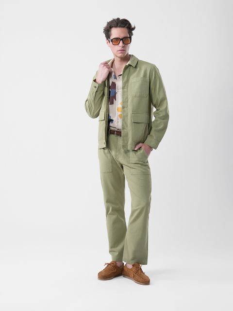 Un hombre que lleva gafas de sol, una chaqueta y pantalones de color verde claro, una camisa estampada y zapatos marrones, está de pie delante de un sencillo fondo blanco.