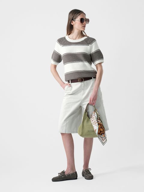 Mujer con gafas de sol que lleva un jersey de rayas de manga corta, pantalones cortos blancos hasta la rodilla y zapatos planos, y que sostiene un bolso verde con una bufanda en la mano.