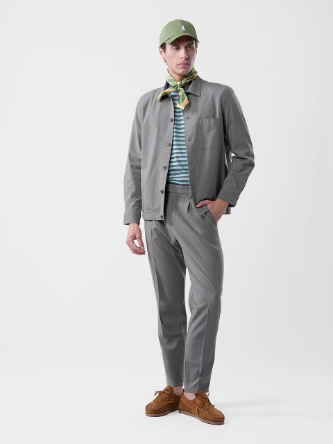 Hombre con traje gris, camisa a rayas, gorro verde, pañuelo estampado y zapatos marrones sobre un fondo blanco liso.