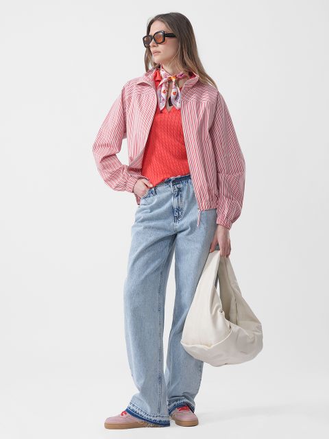 Mujer con gafas de sol lleva una chaqueta de rayas rosas, un jersey rojo, vaqueros azules y una bufanda, y sostiene un bolso blanco grande delante de un fondo blanco liso.