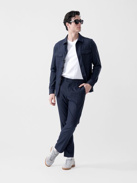 Un hombre que lleva gafas de sol negras, una chaqueta azul marino, una camiseta blanca, pantalones azul marino y zapatillas grises está de pie con un pie cruzado delante del otro y mirando hacia un lado.