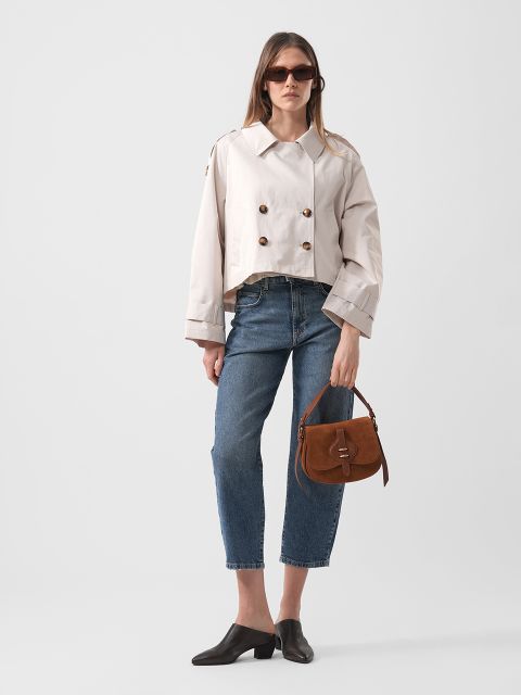 Una mujer con gafas de sol lleva una chaqueta beige de doble botonadura, unos vaqueros azules recortados, zapatos negros y sostiene un bolso marrón delante de un fondo liso.