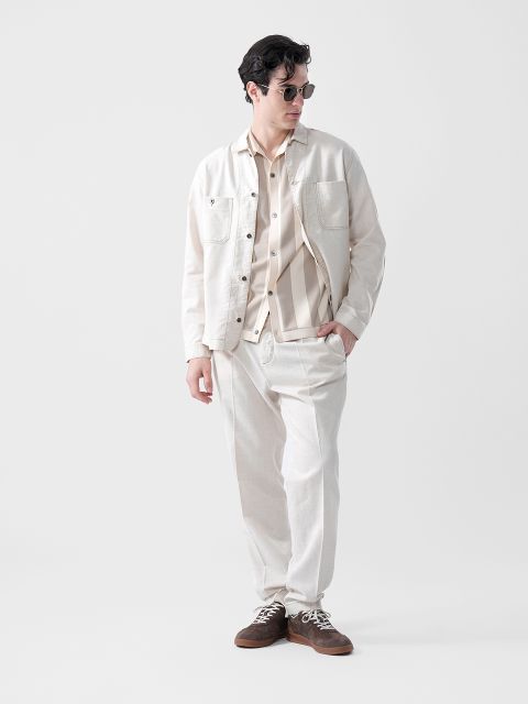 Un hombre con gafas de sol, chaqueta beige claro, camisa a rayas y pantalones a juego está de pie con las manos en los bolsillos frente a un sencillo fondo blanco.