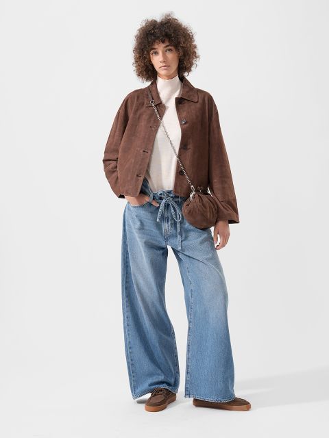 Persona que lleva una chaqueta marrón, una blusa blanca, unos vaqueros azules de corte ancho, zapatos marrones y un pequeño bolso de mano marrón, de pie delante de un fondo liso.