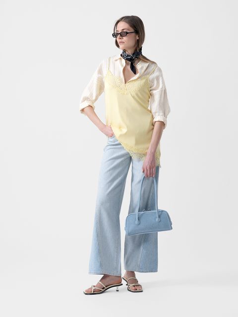Mujer con gafas de sol que lleva una camiseta amarilla sobre una camisa de rayas, vaqueros azul claro de pierna ancha, sandalias y un pañuelo, y que sostiene un bolso azul.