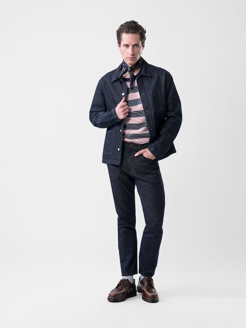 Un hombre posa con una chaqueta y pantalones a rayas azul marino, una camisa a rayas rosa y gris, zapatos marrones y una bufanda estampada contra un fondo blanco liso.