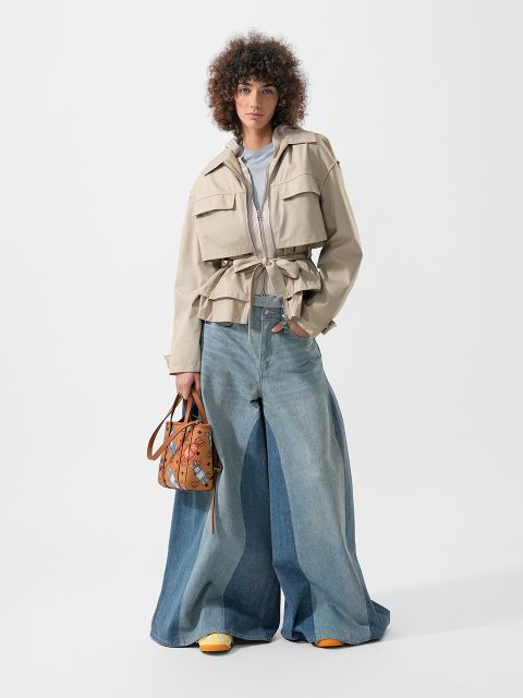 Una persona que lleva una chaqueta beige, vaqueros azules de corte ancho y zapatos amarillos, y que sostiene un bolso de mano con estampado marrón, está de pie delante de un fondo blanco.