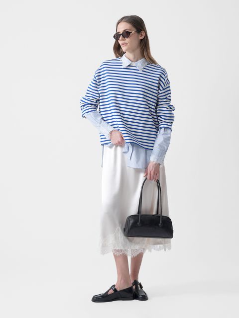 Mujer con gafas de sol que lleva un jersey de rayas azules y blancas sobre una camisa con cuello, una falda blanca con adornos de encaje, zapatos negros y un bolso negro.