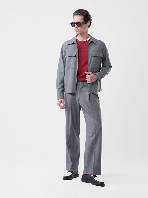Hombre con gafas de sol, traje gris, camisa roja, zapatos blancos y negros y un collar, sobre un fondo blanco.