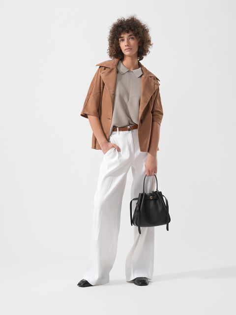 Mujer con cabello rizado que lleva una chaqueta marrón claro, una camisa beige y pantalones blancos de pierna ancha, sosteniendo un bolso negro, de pie frente a un fondo sencillo.