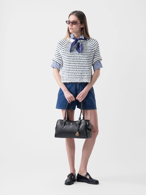 Mujer con gafas de sol que lleva un jersey de rayas sobre una camisa con cuello, pantalones cortos vaqueros y zapatos negros, y que sostiene un bolso negro delante de un fondo blanco.