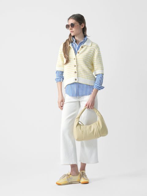 Mujer con gafas de sol, camisa azul a rayas, cárdigan de punto a rayas, pantalón blanco y zapatos amarillos sostiene un bolso de mano amarillo claro y está de pie frente a un fondo blanco.
