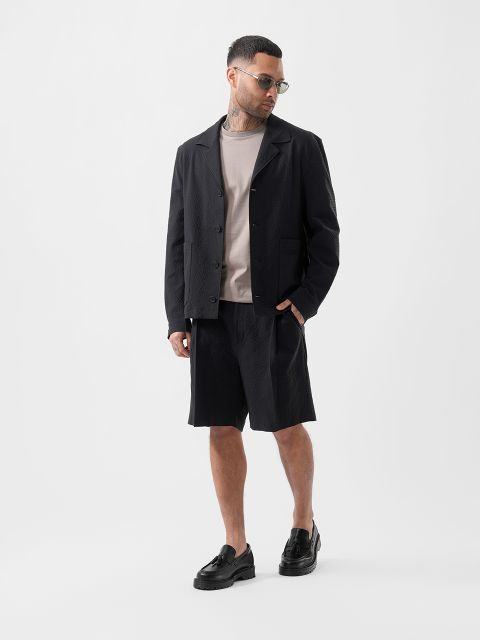 Un hombre con gafas de sol, chaqueta negra texturizada, camiseta beige, pantalones cortos negros y zapatillas negras está de pie frente a un fondo blanco liso.