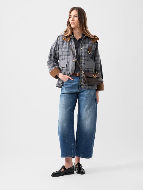 Una mujer con una chaqueta a cuadros, jeans azules de pierna ancha, zapatos negros y un bolso bandolera marrón está de pie frente a un fondo blanco liso.