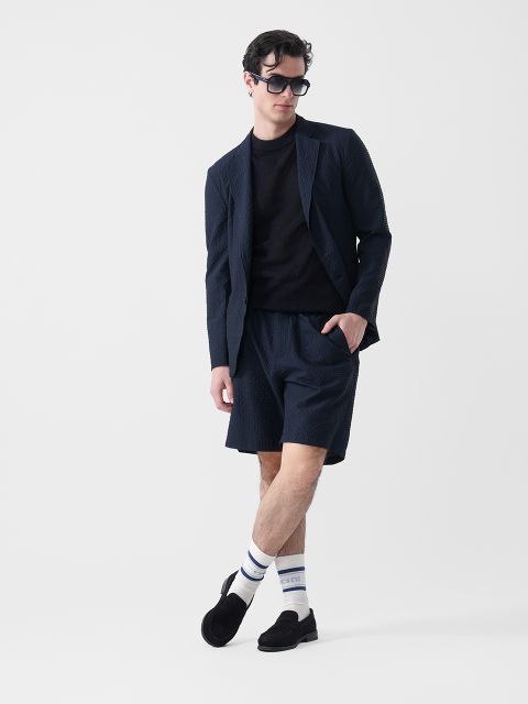 Un hombre que lleva un blazer oscuro, pantalones cortos a juego, una camiseta negra, gafas de sol, calcetines blancos con rayas y mocasines negros está de pie delante de un fondo blanco.