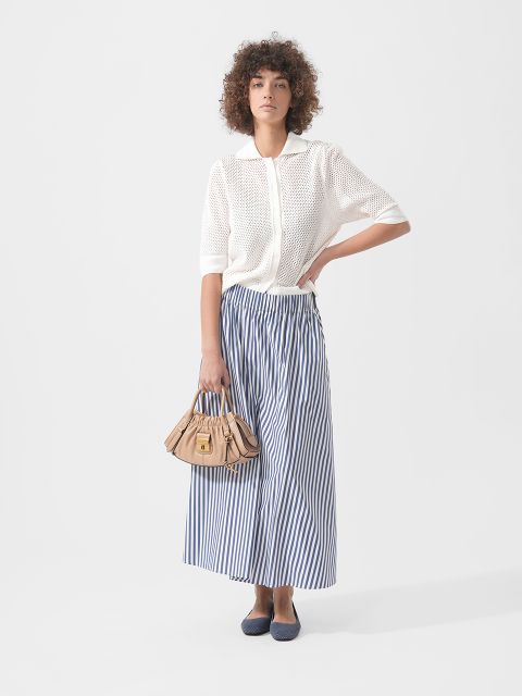 Mujer con un top de punto blanco de manga corta, una falda maxi a rayas azules y blancas, zapatos planos azul marino y un bolso beige delante de un fondo liso.