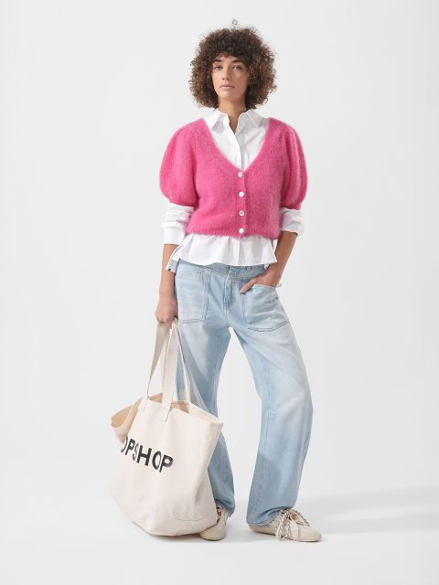 Una persona con una chaqueta de punto rosa, camisa blanca, vaqueros azul claro y zapatillas de deporte sostiene una gran bolsa de mano beige con la inscripción "TOPSHOP".