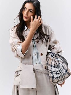 Mujer con ropa de color beige claro y un bolso de cuadros, mirando hacia un lado y tocándose el cabello frente a un fondo sencillo.