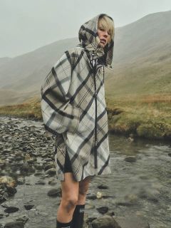 Una persona que lleva un impermeable a cuadros con capucha está de pie junto a un arroyo rocoso en un paisaje neblinoso y cubierto de hierba, con colinas al fondo.