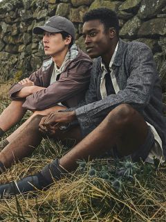 Dos hombres sentados con ropa informal de exterior sobre hierba seca junto a un muro de piedra, mirando pensativos a lo lejos.