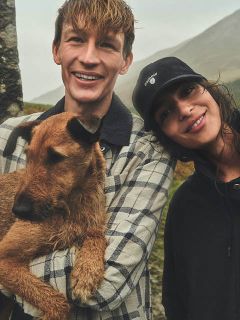 Un hombre sonriente sostiene un perro marrón mientras está de pie junto a una mujer sonriente con un gorro negro, al aire libre en un día nublado.