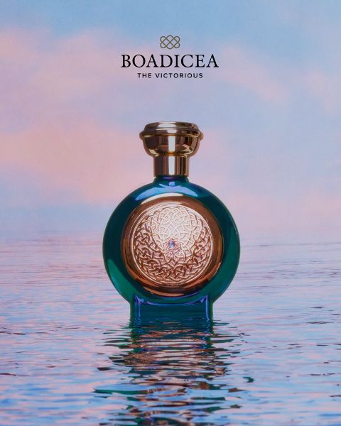 Un frasco de perfume azul y dorado de «Boadicea» reposa sobre agua lisa como un espejo, con el telón de fondo de un cielo de color pastel.