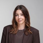 Una mujer con cabello castaño ondulado, que viste un blazer marrón y collares de varias capas, posa frente a un fondo gris claro liso.