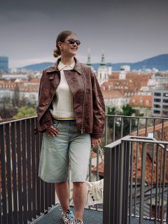 Mujer con gafas de sol y chaqueta marrón de pie en un balcón con vistas a un paisaje urbano con tejados rojos y torres de iglesias en un día nublado.