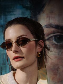 Mujer de cabello castaño, que lleva gafas de sol ovales y un pendiente dorado, de pie frente a un retrato pintado con un ojo grande y tonos azules.
