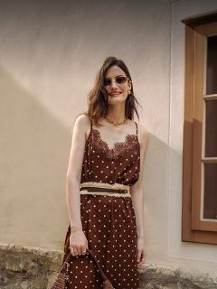Una mujer con un vestido marrón de lunares con adornos de encaje, gafas de sol y joyas de oro está de pie sonriendo frente a una pared beige y una ventana.