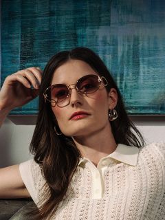Mujer con cabello largo castaño, que lleva gafas de sol, pendientes y una parte superior blanca con cuello, sentada frente a una pintura abstracta azul.