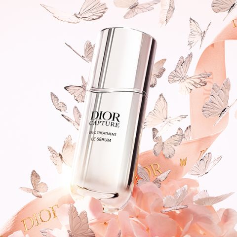 Der Flakon von Dior Capture Le Sérum ist umgeben von rosa Blütenblättern, zarten Schmetterlingen und einem Band mit dem Text "Dior" auf einem sanften pastellfarbenen Hintergrund.