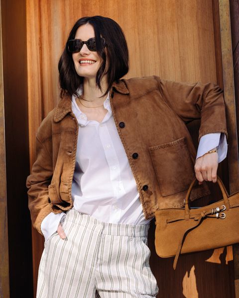 Mujer con gafas de sol, que lleva una chaqueta marrón, una camisa blanca y pantalones a rayas, sonríe y sostiene un bolso marrón delante de un fondo de madera.