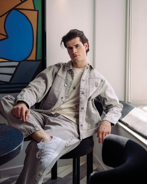 Un joven con una chaqueta vaquera clara y zapatillas blancas está sentado relajado en una silla junto a la ventana en una habitación moderna con arte abstracto en la pared.