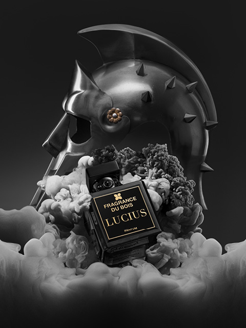 En medio del humo, hay un frasco de perfume negro con la inscripción «Fragrance Du Bois Lucius»; en el fondo se ve un casco de gladiador metálico.