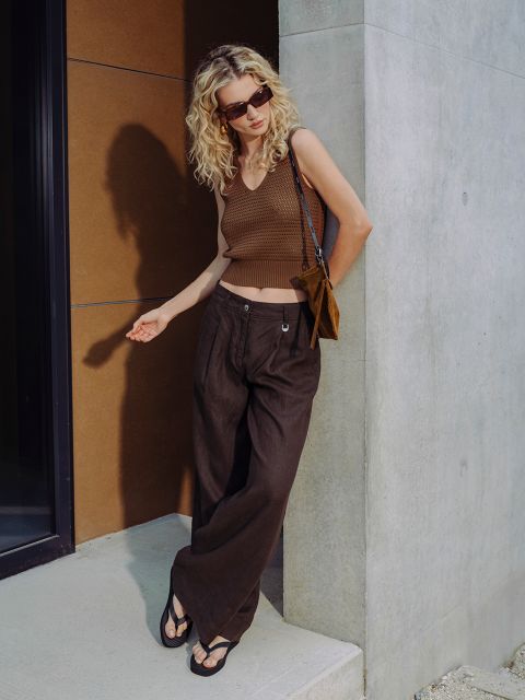 Una mujer con cabello rubio rizado lleva gafas de sol, un top sin mangas marrón, pantalones anchos marrones y sandalias, y está de pie frente a un edificio moderno.